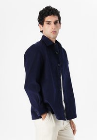 Camisa de pana azul marino con cierre de botones en la parte delantera, cuello puntiagudo, bolsillo en el pecho y mangas largas con puños abotonados. Textura suave.