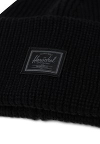 Bonnet en tricot noir avec une texture côtelée et une étiquette noire rectangulaire portant des lettres blanches mettant en avant la marque Herschel.