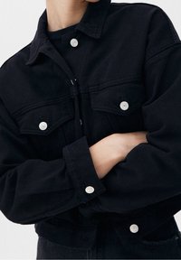 Zwarte spijkerjas met zilveren knopen, twee borstzakken en een losse pasvorm. Geweven stof met een kraag en lange mouwen.