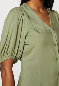Olivgrüne Bluse mit puffierten kurzen Ärmeln, V-Ausschnitt und Knopfdetail entlang der Vorderseite, aus einem glatten, leicht glänzenden Stoff.