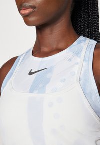 Bela športna majica brez rokavov s svetlo modrim akvarelnim vzorcem, okroglim izrezom in črnim logotipom Nike na zgornjem srednjem delu.