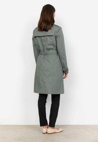 Frau trägt einen grauen Trenchcoat mit Gürtel, schwarze enge Hosen und beige flache Schuhe, steht mit dem Rücken zur Kamera vor weißem Hintergrund.