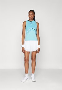 Top atlético sin mangas de color azul claro con ribetes negros y diseño abstracto, combinado con una falda de tenis blanca con pliegues y zapatillas blancas.