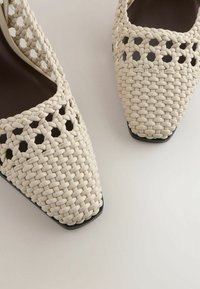 Chaussures beiges tissées à bout pointu avec un design en treillis ouvert et une semelle intérieure noire, photographiées sur une surface blanche.