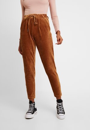 Persoon draagt een broek van bruin ribfluweel met hoge taille en trekkoord, een roze geribd lang mouw shirt en zwarte sneakers met hoge bovenkant.