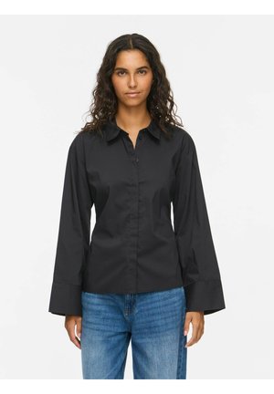 Femme aux cheveux bruns ondulés portant une chemise noire à manches longues avec de larges poignets et un jean bleu, debout devant un fond blanc uni.