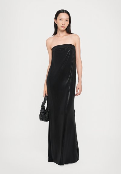 Woman wearing a strapless, floor-length black satin dress holding a small black handbag with a wide strap against a plain white background.