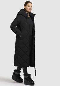 Sort lang dunjakke med diamantquiltet design, oversized hætte, lynlåslukning og sidelommer. Kombineret med sorte snørebåndsstøvler.