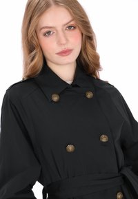 Zwarte trenchcoat met een dubbelgeknoopt ontwerp, voorzien van gouden knoppen en een tailleband. Gladde stof met een getailleerde pasvorm.