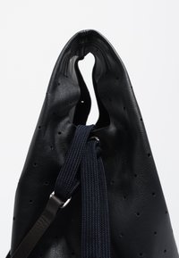 Czarna skórzana torba z perforacjami, o luźnym kształcie i teksturowanym wykończeniu. Posiada granatowe zapięcie na sznurek oraz metalowe akcenty.