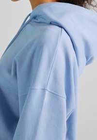 Sudadera con capucha de algodón azul claro con cordón, que presenta un ajuste relajado, textura suave y detalles visibles de costura en la costura del hombro.