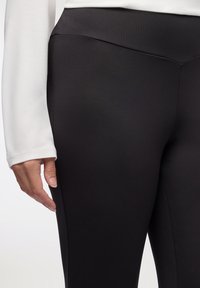 Czarne legginsy wykonane z gładkiego, elastycznego materiału, z wysokim stanem i płaskimi szwami; ręka opiera się na boku dla podparcia.