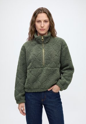 Jeune femme aux cheveux ondulés portant un pull polaire vert texturé avec une fermeture éclair dorée et un jean bleu, debout sur un fond uni.