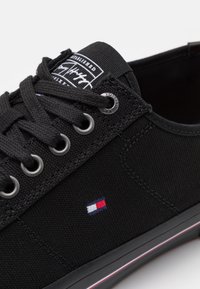 Baskets en toile noire avec des lacets plats noirs, des œillets argentés et une semelle en caoutchouc rayée. Comprend un patch de logo et un petit emblème coloré.