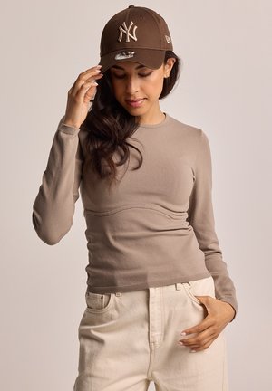 Beige Langarmoberteil mit einem taillierten Design und einer geschwungenen Naht an der Vorderseite, kombiniert mit cremefarbigen Hosen und einer braunen Baseballkappe.