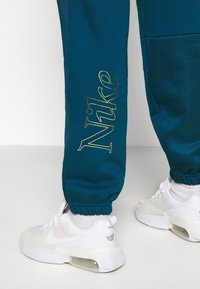 Pantalones jogger en color verde azulado con el logo dorado de "Nike" y cinturilla elástica. Combinados con zapatillas blancas y detalle de amortiguación de aire visible.