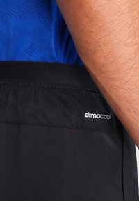 Närbild av en persons midja som bär svarta Climacool-shorts och en blåmönstrad skjorta, som visar en del av deras arm och kläddetaljer.