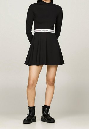 Woman wearing a black long-sleeve dress with a white "Tommy Jeans" belt and black lace-up combat boots, standing against a plain background.