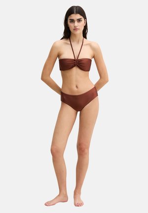 Młoda kobieta stoi boso, ubrana w brązowy, bezramiankowy top bikini z ramiączkami wiązanymi na szyi oraz pasujące do niego bikini z wysokim stanem.