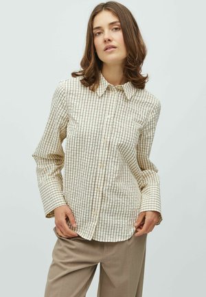 Mlada ženska, oblečena v bež srajco s karirastim vzorcem in svetlo rjave hlače, stoji pred belim ozadjem.