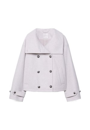 Femme portant une veste blanche à col montant sur un top court rayé, un pantalon noir ample et des bottes noires montantes sur un fond gris uni.