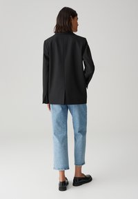 Zwarte op maat gemaakte blazer, ontspannen fit, met een split aan de achterkant. Gecombineerd met blauwe denim rechte spijkerbroek en zwarte instap schoenen.
