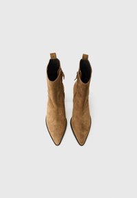 Bottines en suède marron avec des bouts pointus et des zips sur les côtés, posées sur un fond gris clair.
