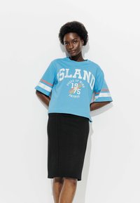 Femme aux cheveux courts et bouclés portant un T-shirt graphique bleu et une jupe noire mi-longue, se tenant devant un fond blanc.