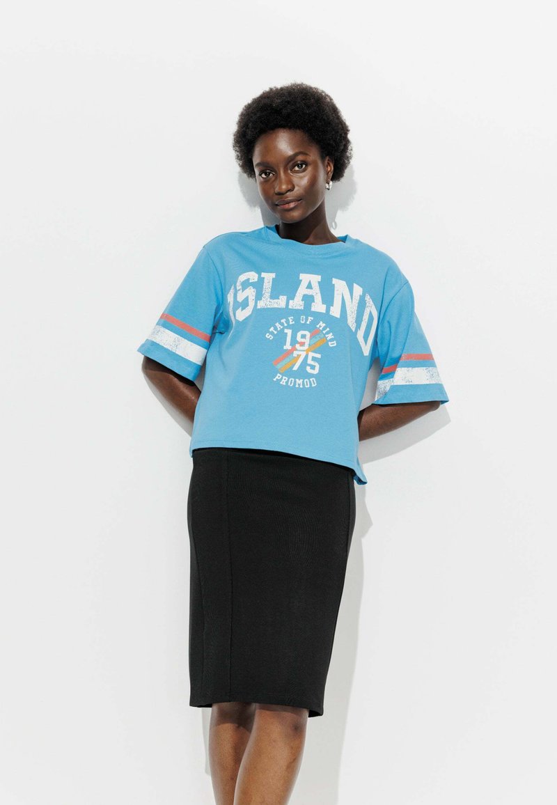 Femme aux cheveux courts et bouclés portant un T-shirt graphique bleu et une jupe noire mi-longue, se tenant devant un fond blanc.