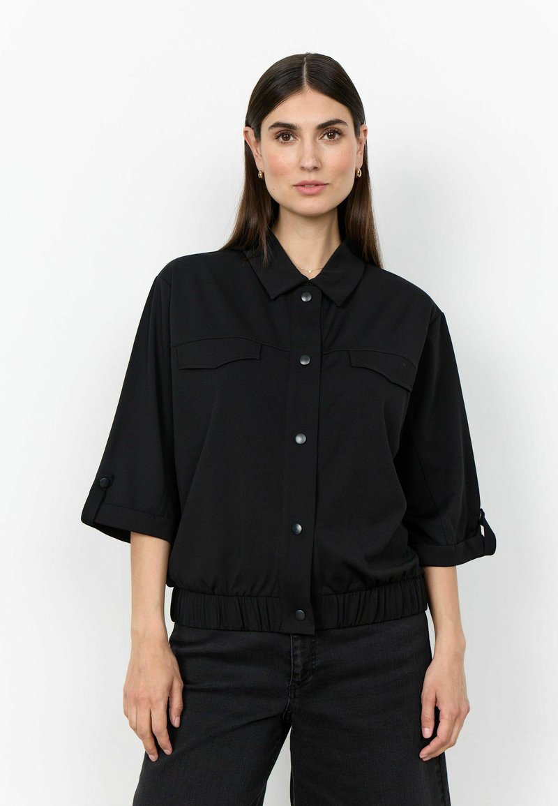 Femme aux cheveux raides et foncés portant une chemise noire boutonnée avec des manches trois-quarts et un pantalon noir, debout sur un fond blanc.