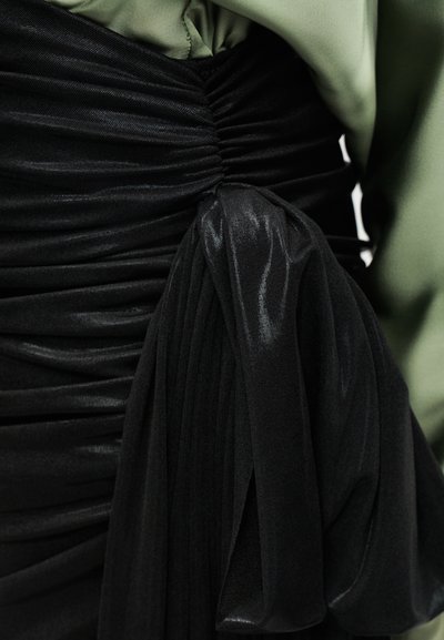 Κοντινό πλάνο μαύρου υφάσματος με ρίγες, συγκεντρωμένο στη μέση με μεγάλο δεμένο φιόγκο, φορεμένο με ένα ελιά πράσινο μακρυμάνικο ένδυμα.