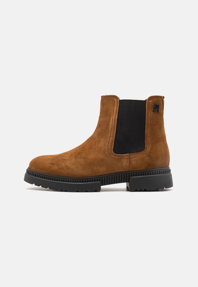 Botte Chelsea en daim marron avec panneau latéral élastique noir, semelle en caoutchouc noir et petit logo noir près de la cheville sur un fond clair uni.
