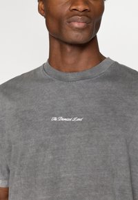 Γκρι βαμβακερό μπλουζάκι με στρογγυλή λαιμόκοψη, που διαθέτει λευκό καλλιγραφικό κείμενο "Η Υποσχεμένη Γη" στο μπροστινό μέρος. Μαλακή υφή με ξεθωριασμένο σχέδιο.