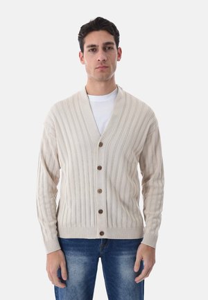 Uomo che indossa un cardigan beige con bottoni sopra una t-shirt bianca e jeans blu, in piedi rivolto verso l'osservatore su uno sfondo semplice.