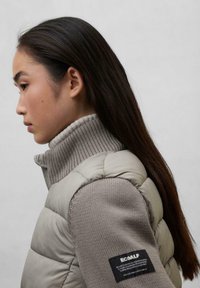 Gilet leggero e imbottito di colore beige con dolcevita grigio a coste. Presenta un'etichetta nera sulla manica. Texture liscia e design aderente.