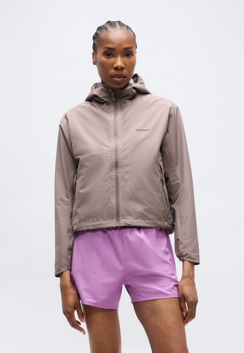 Femme aux cheveux tressés portant une veste à capuche beige zippée et un short violet clair, debout devant un fond clair uni.