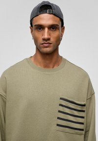 Olijfgroen sweatshirt met een ronde halslijn en een zwart gestreept zakdetail. Grijze pet achterstevoren gedragen. Zachte textuur, casual ontwerp.