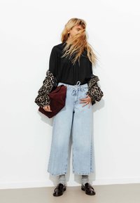Jean en denim bleu clair à jambes larges, associé à un haut noir et des manches imprimées léopard, tenant un sac en daim bordeaux. Mocassins noirs et chaussettes grises.