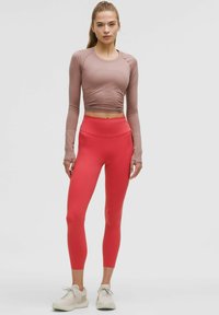 Długi rękaw, dopasowany, jasnoróżowy crop top zestawiony z wysokim stanem, elastycznymi, czerwonymi legginsami. Dodatki to jasne buty sportowe.