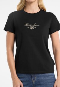Must musta T-särk, millel on kuldne tikand "Guess Jeans" logo koos ümmarguse embleemiga, ümmargune kaelus ja lühikesed varrukad.