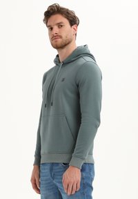 Man met kort bruin haar en baard draagt een gedempt groene hoodie en blauwe jeans, staand tegen een effen witte achtergrond.