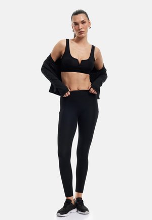 Vrouw draagt zwarte sportbeha, zwarte legging en zwarte sneakers, past de tailleband aan, jas glijdt van haar schouders, staand tegen een witte achtergrond.