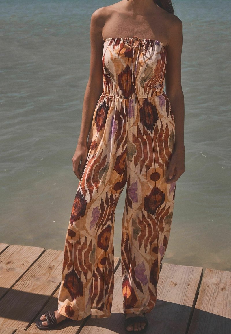 Femme portant une combinaison bustier à motifs avec des jambes larges, debout sur un quai en bois près de l'eau.