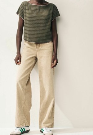 Personne portant un haut ample à rayures vert olive, un jean beige à jambes larges, et des baskets blanches avec des rayures vertes, debout contre un mur uni.