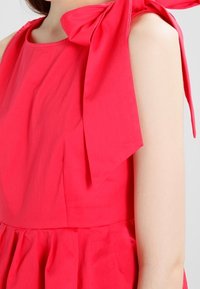 Fuksjowa sukienka z okrągłym dekoltem, marszczoną talią i dużą kokardą na ramieniu, wykonana z lekkiego materiału o gładkiej fakturze.