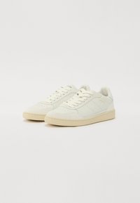 Hvite skinn sneakers med perforert design, flate lisser, kremfarget gummisåle og subtil merkevare på siden. Jevn tekstur med aksenter.