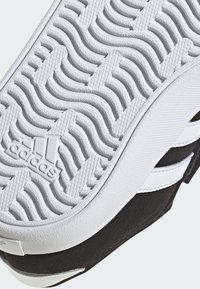 Černobílá podrážka tenisky s texturovaným bílým gumovým vzorem pro lepší přilnavost, která obsahuje vyražené logo Adidas a tři bílé pruhy.