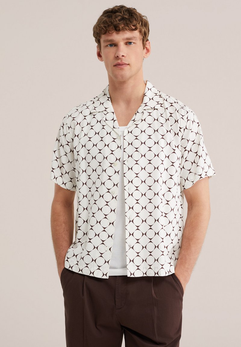 Junger Mann mit lockigem Haar, der ein kurzärmeliges weißes Hemd mit schwarzem geometrischem Kreis-Muster und braune Hose trägt, die Hände in den Taschen.