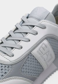 Αθλητικό παπούτσι Emporio Armani σε ανοιχτό γκρι και λευκό, με υφή από πλεκτό δίχτυ, με επωνυμία και κορδόνια ιδίου χρώματος σε κοντινή λήψη.