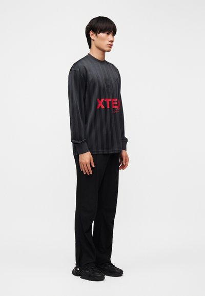 Νεαρός άνδρας που φοράει μαύρο ριγέ μακρυμάνικο πουκάμισο με κόκκινο κείμενο, μαύρο παντελόνι και μαύρα αθλητικά παπούτσια, στέκεται μπροστά σε λευκό φόντο.